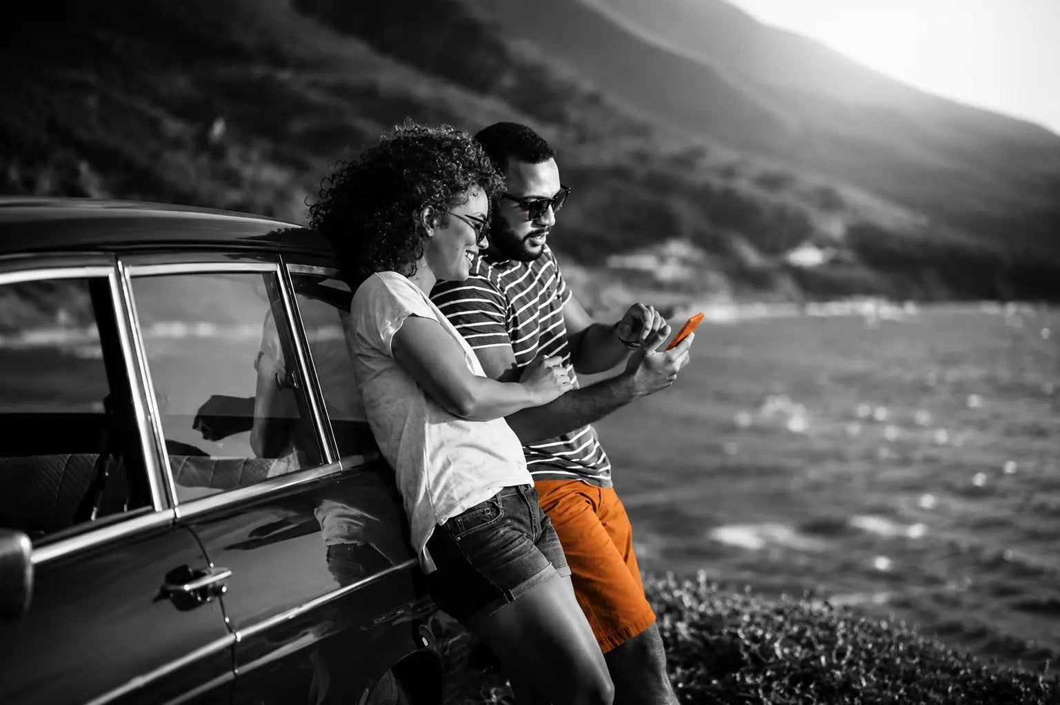 two people lean on a car while looking at their phone near a body of water